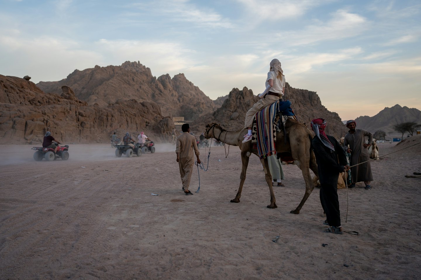 People ride camels and quad bikes across a desert landscape.